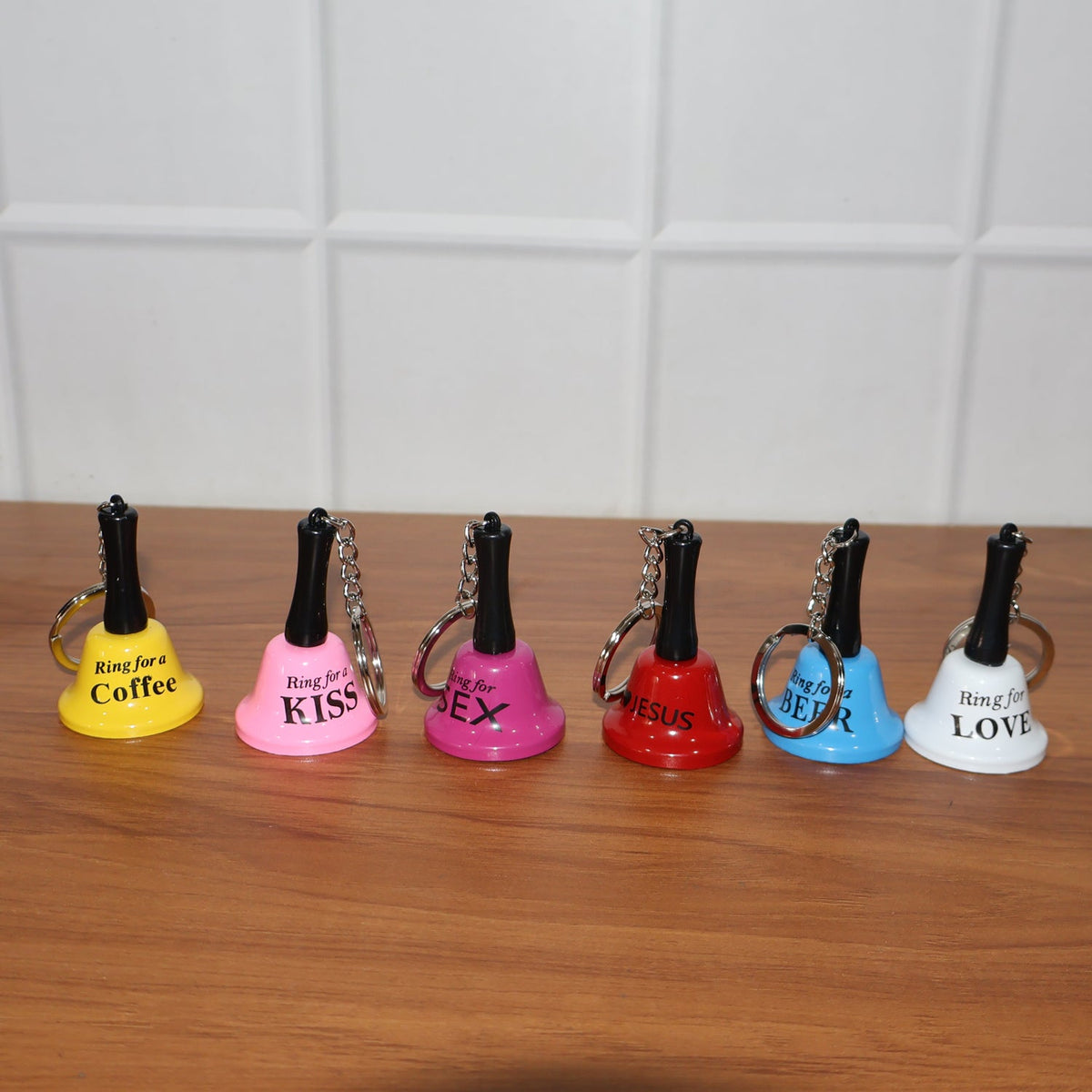 Six mini hand bell keychains in different colors, each with a unique phrase, lined up on a dark wooden surface.