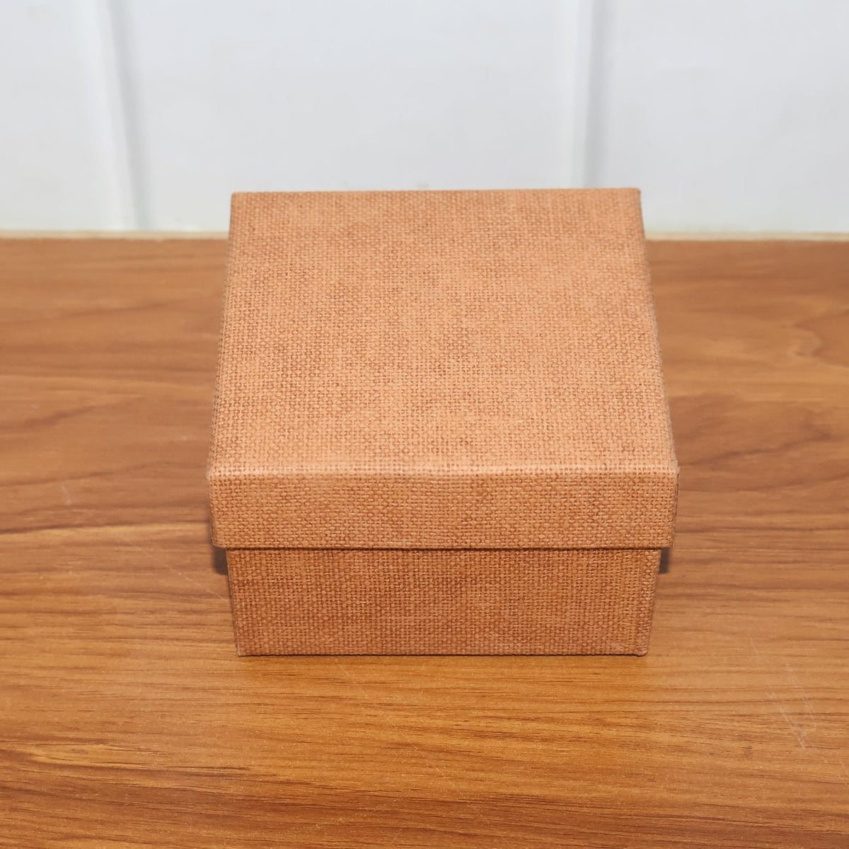 A closed, square, brown textured gift box resting on a light brown wooden surface.