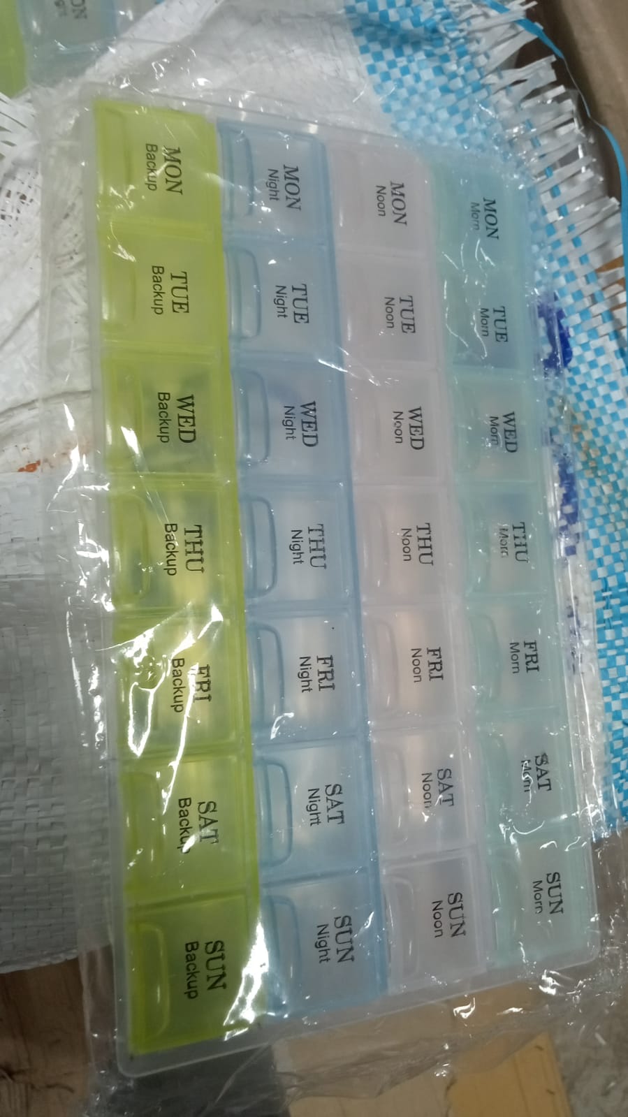 A 28-slot pill organizer, partially wrapped in plastic, with green and blue sections visible.