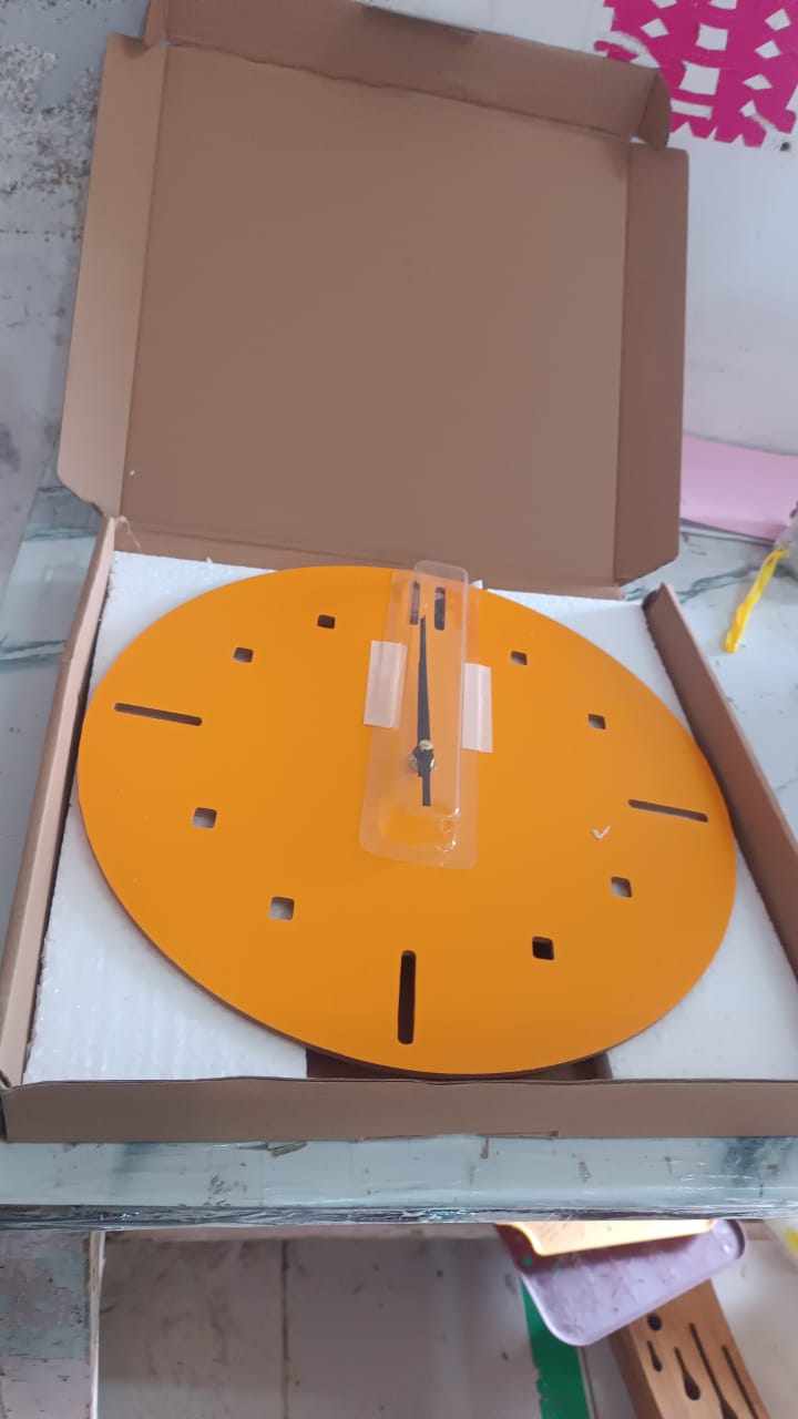A yellow round clock face with black hands and hour markers is packaged in a brown cardboard box with white foam padding.