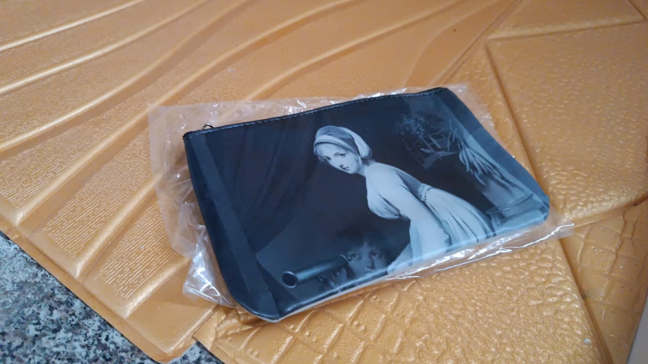 A hand holding a black travel pouch with a classic painting, next to an iPhone and makeup items.