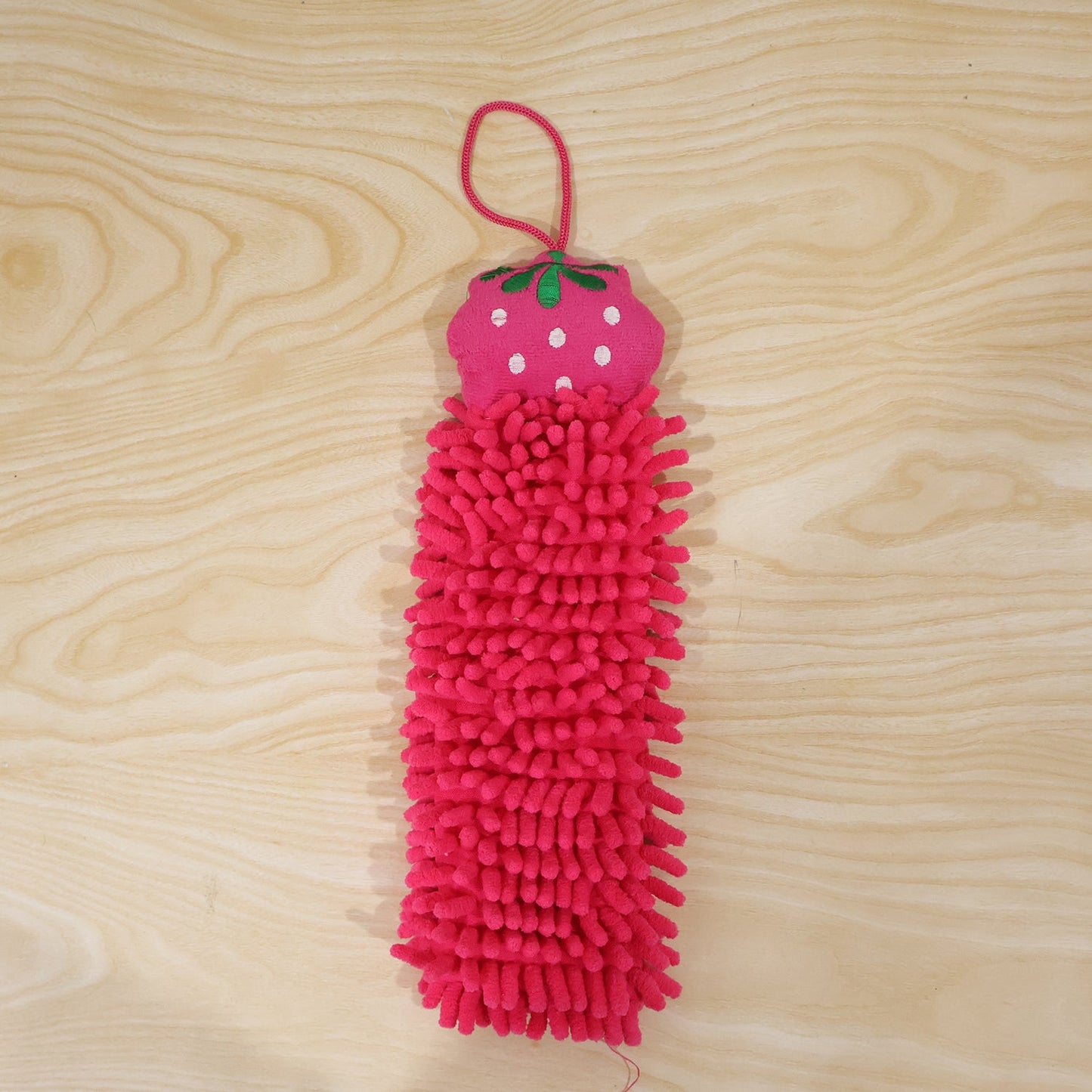 A pink microfiber hand towel with a strawberry top and hanging loop is displayed on a light wooden background.