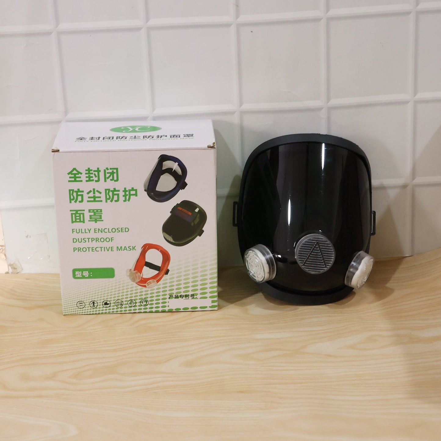 Black full face respirator mask next to its white and green product box on a light wooden surface.