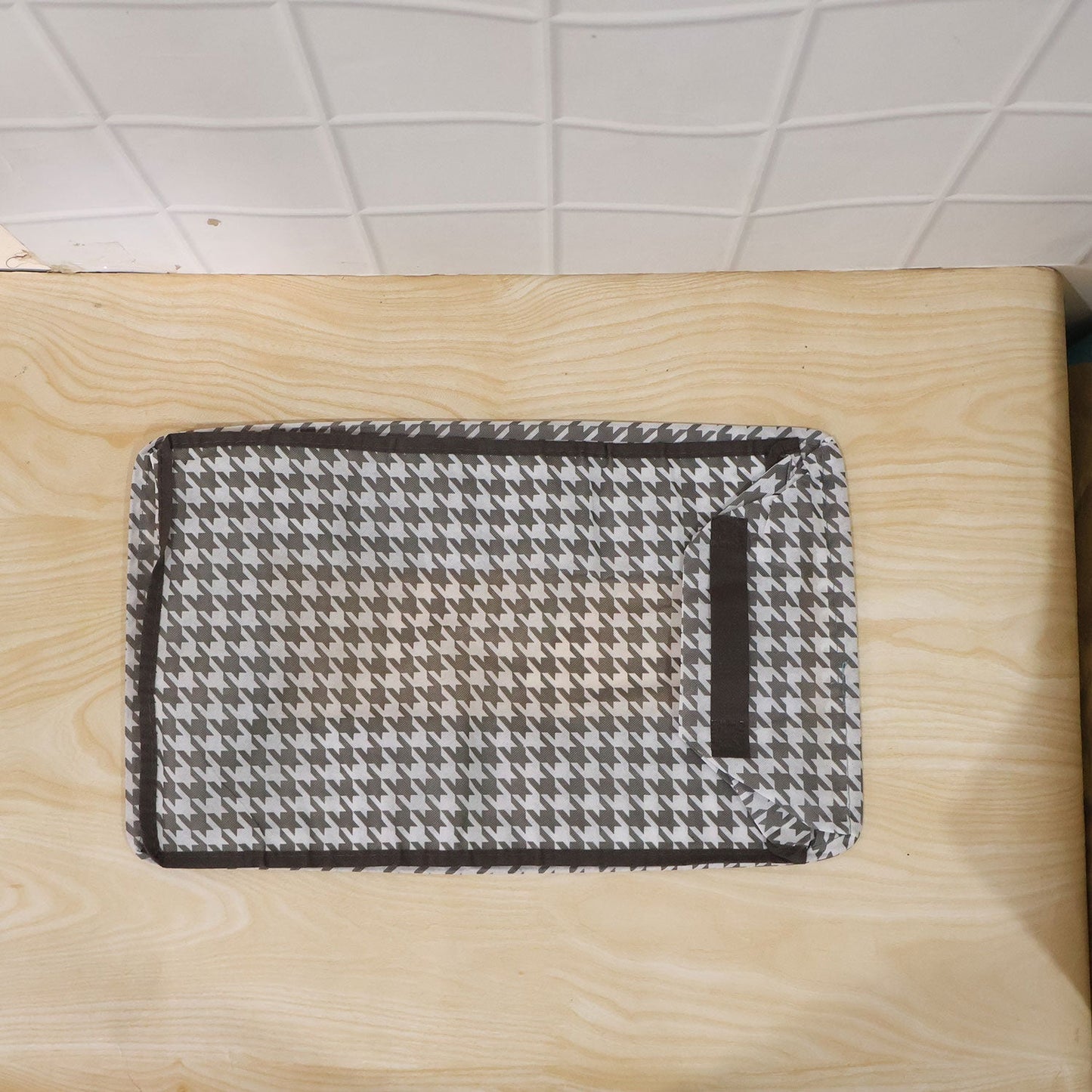 A gray and white houndstooth fabric organizer lies flat on a light wood surface, with a dark brown border.