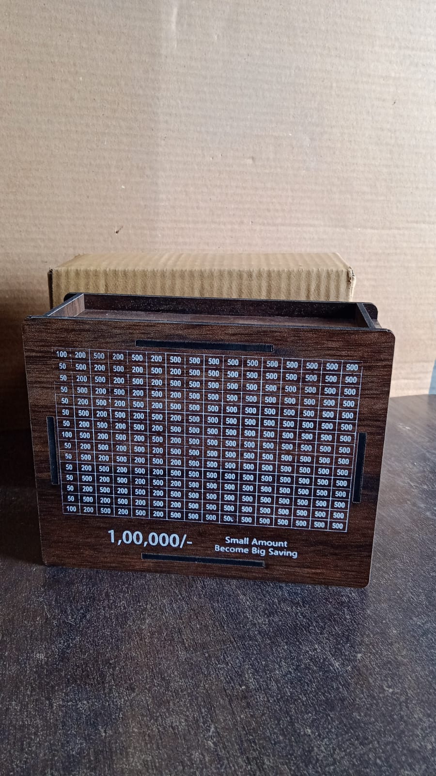 A dark brown wooden savings box with a white grid tracker on a dark surface, with a cardboard box in the background.