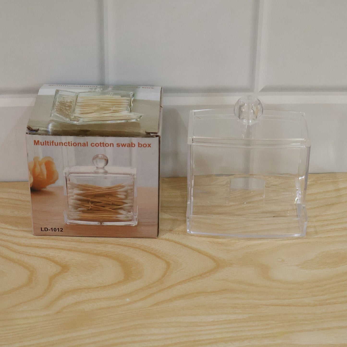 Clear acrylic cotton swab dispenser next to its product packaging box on a wooden surface.