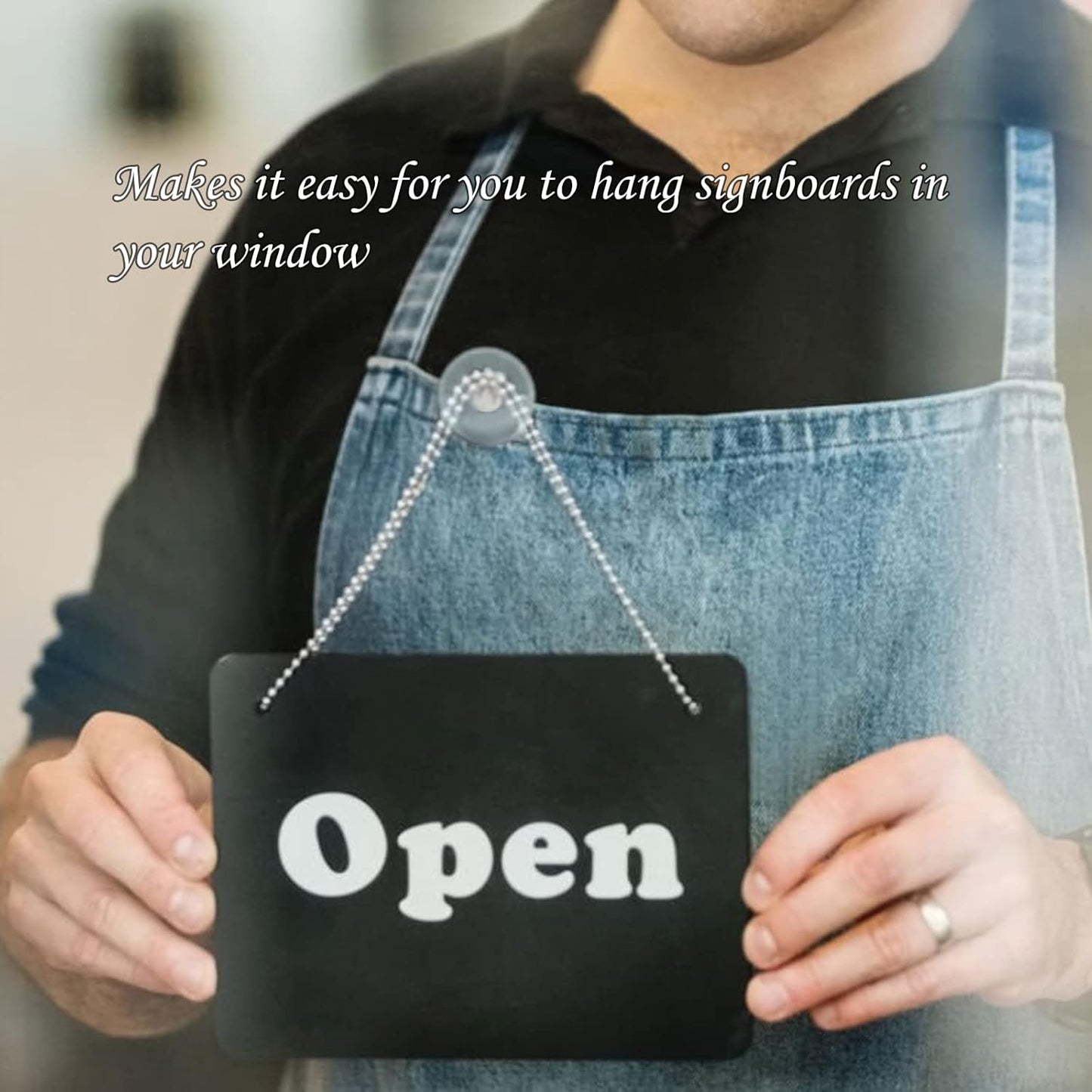 A person in a denim apron holds a black 'Open' sign, attached by a chain to a suction cup hook on a window.