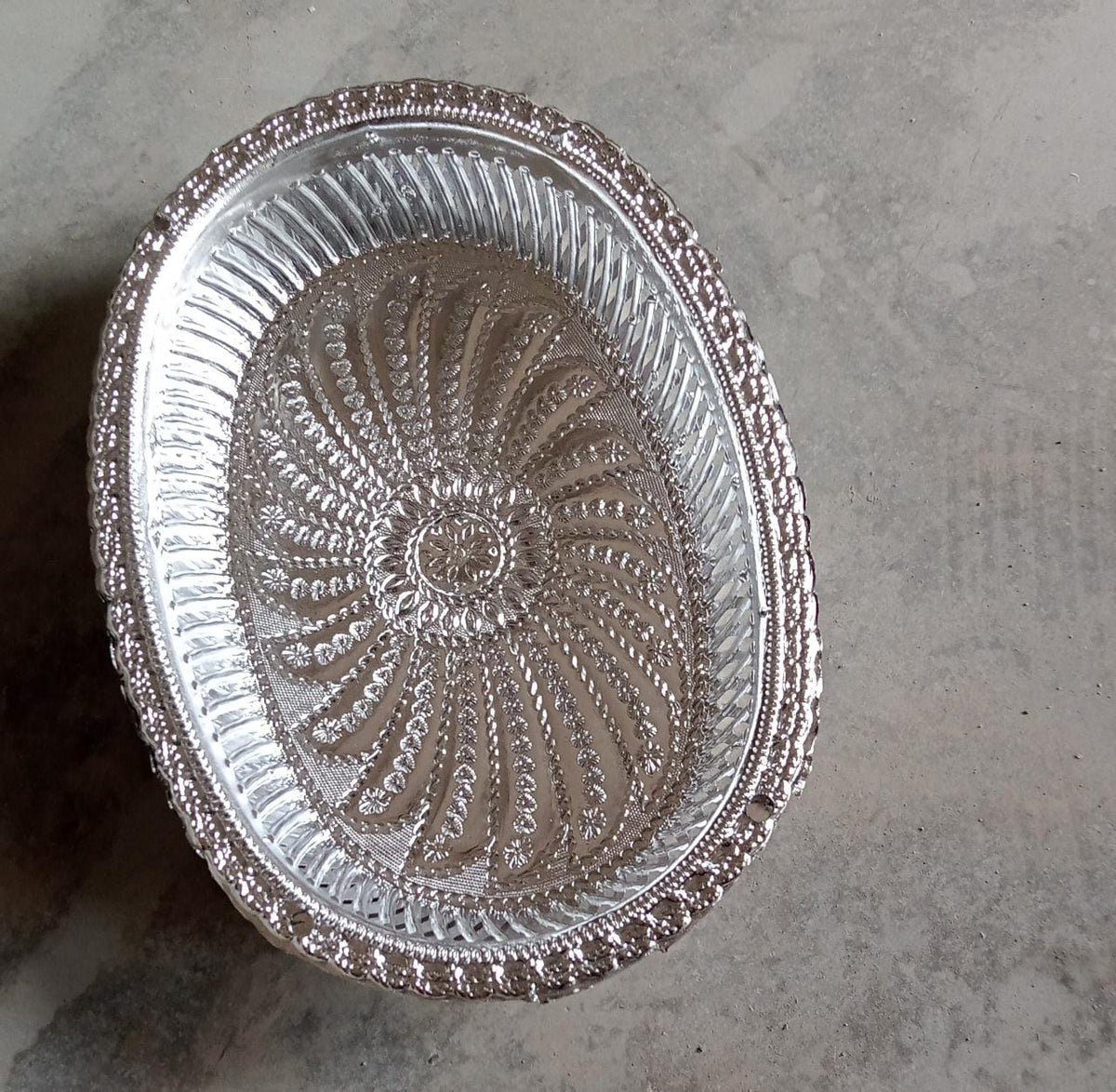 A silver oval decorative serving tray with a floral pattern on a grey concrete background.