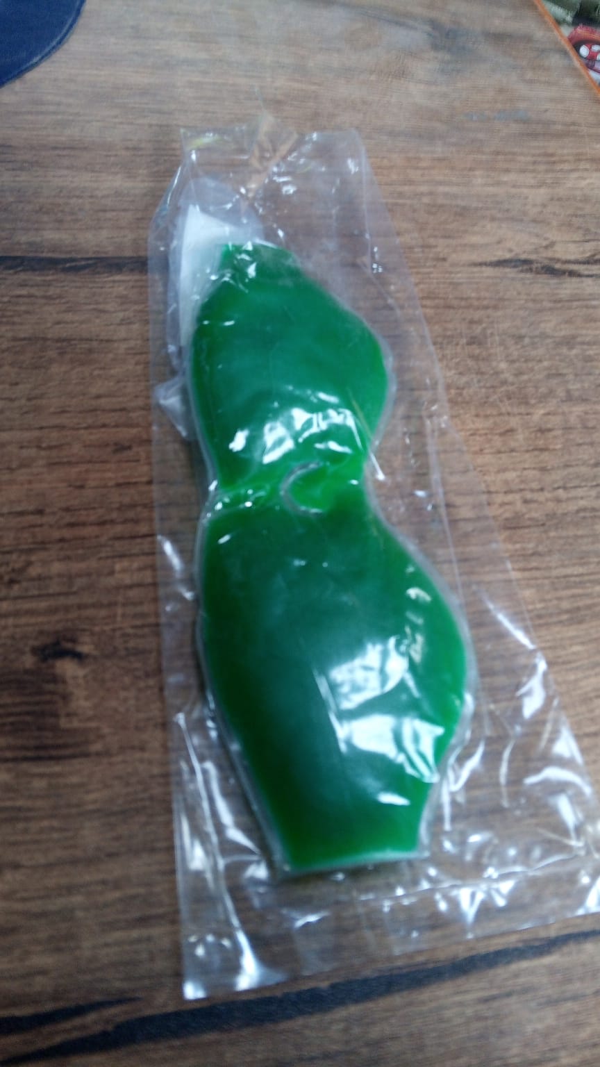A green gel eye mask is sealed in clear plastic packaging, resting on a wooden surface.