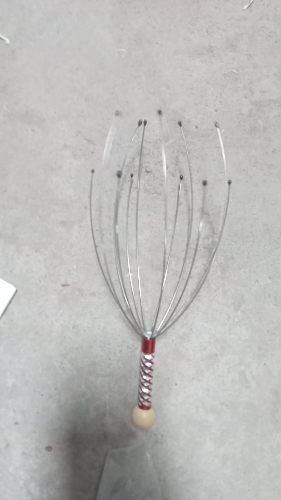 A metal scalp massager with a red and white handle and wooden top is placed on a textured gray surface.