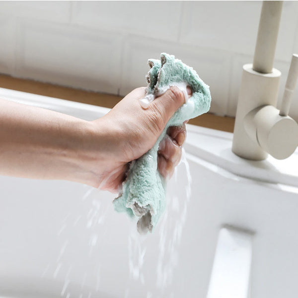 A hand firmly wrings out a light green microfiber towel under running water in a white kitchen sink.