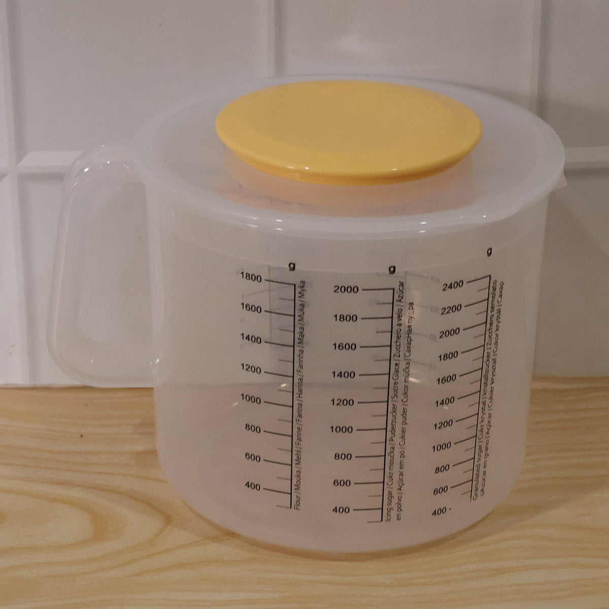 A clear plastic measuring jug with a yellow lid, placed on a light wooden surface against a white tiled wall.
