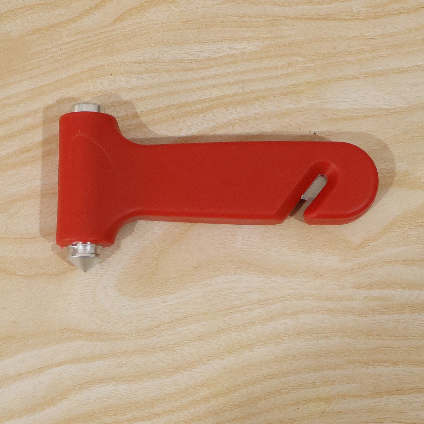 A red car safety hammer with a silver window breaker and seatbelt cutter is placed on a light wood surface.