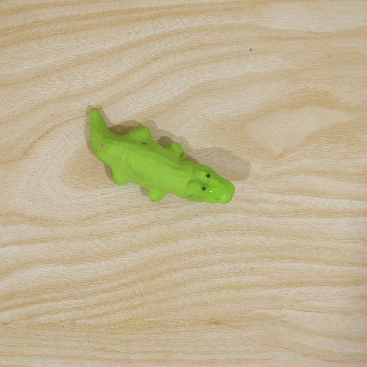 A single green crocodile-shaped eraser is positioned on a light wooden surface with visible grain.