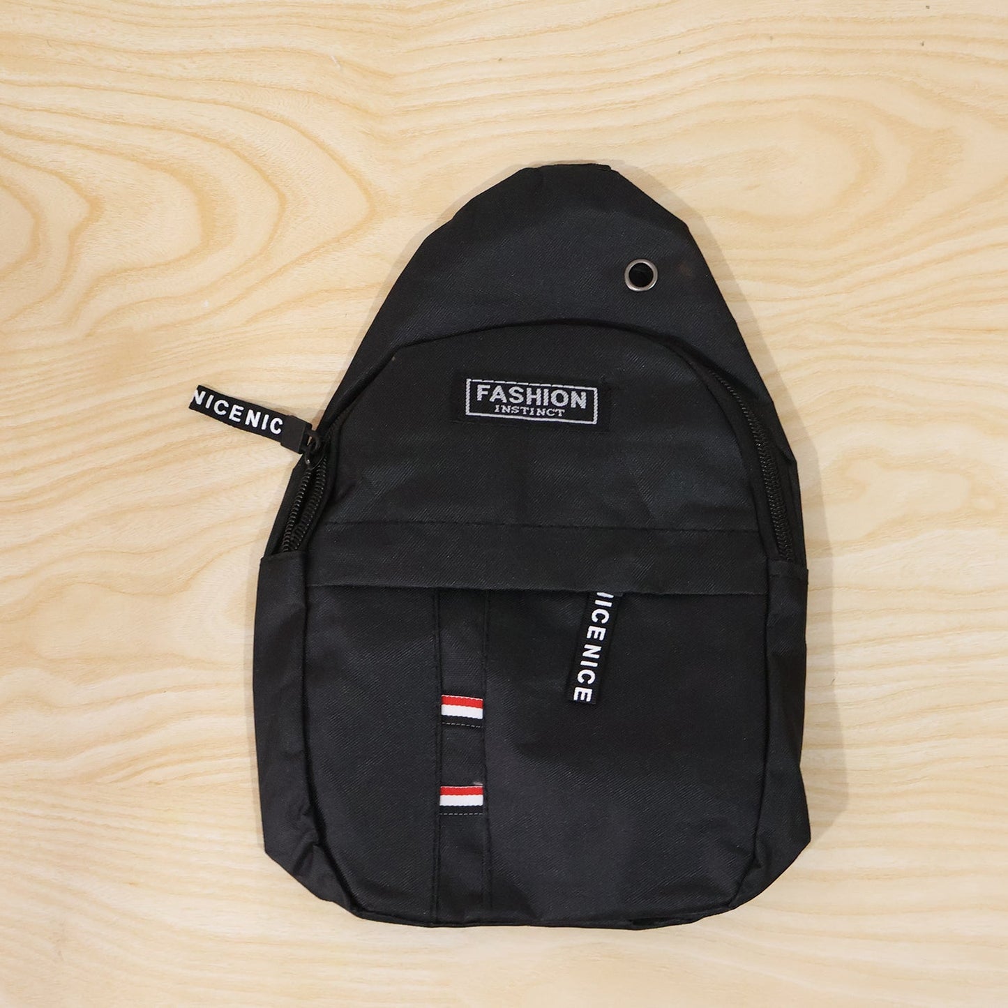 A black sling bag with a front pocket and 'FASHION INSTINCT' label, lying flat on a light wooden surface.