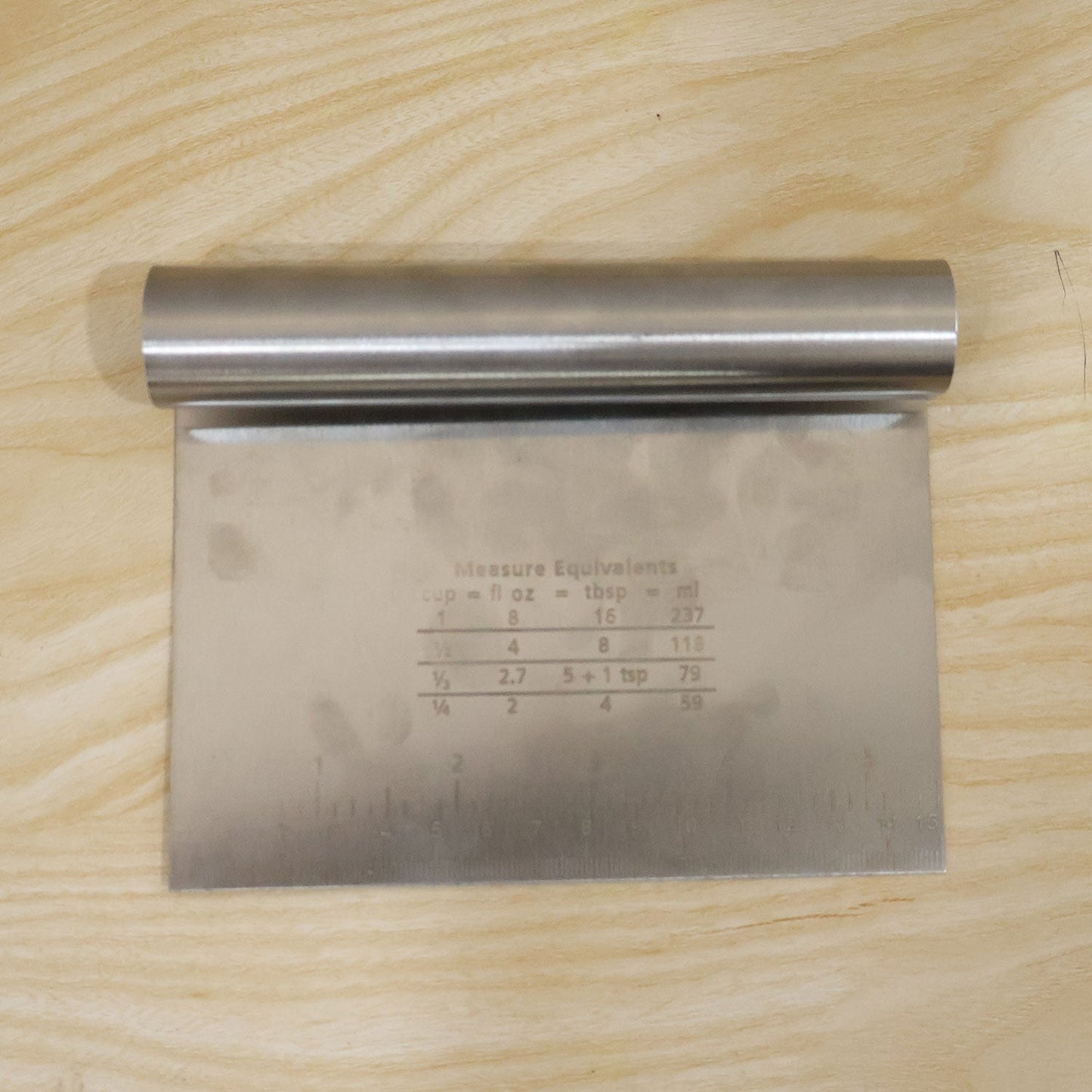 A stainless steel dough scraper with measurement markings and conversion chart sits on a light wooden surface.
