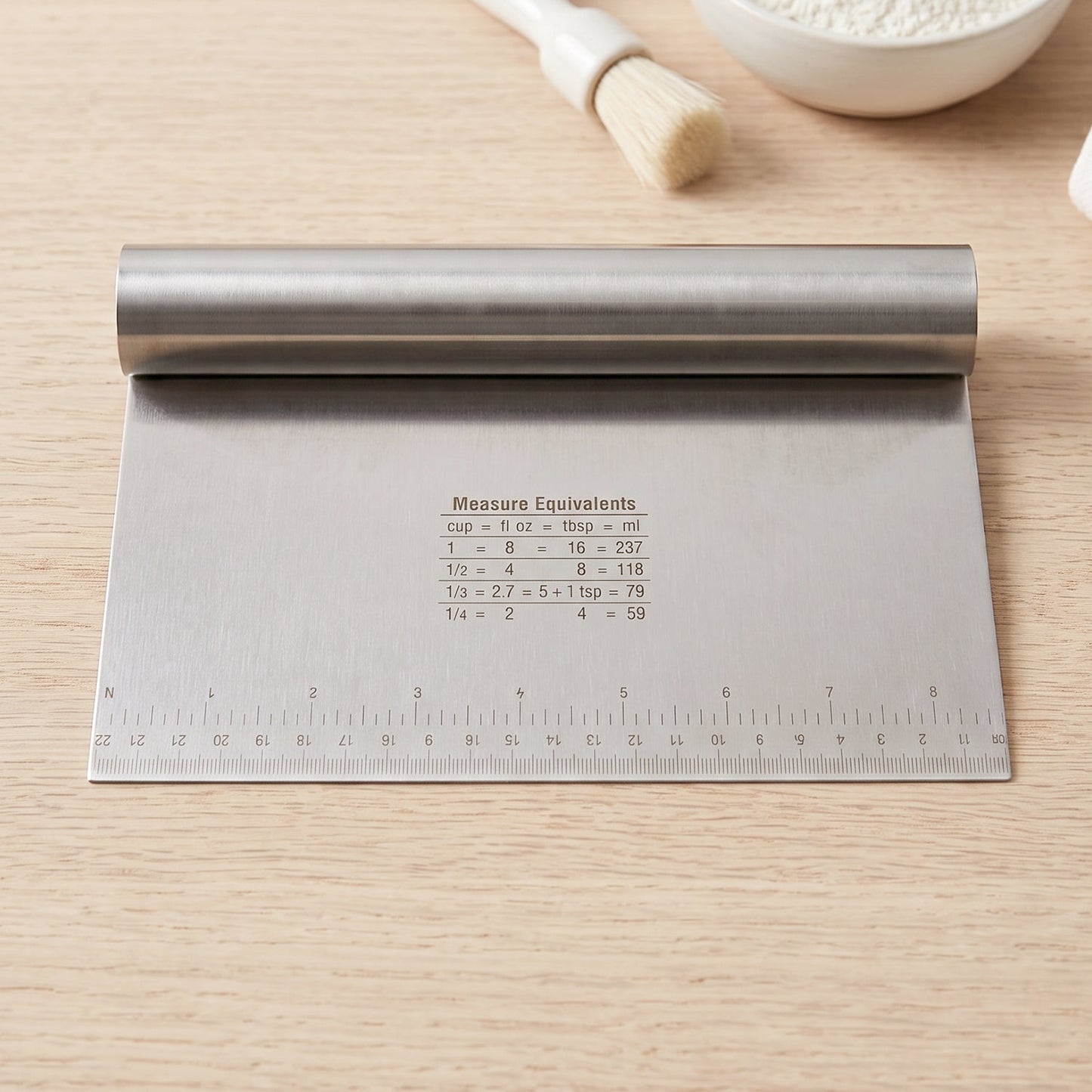 A stainless steel dough scraper with a ruler and measurement equivalents is placed on a light wooden table.