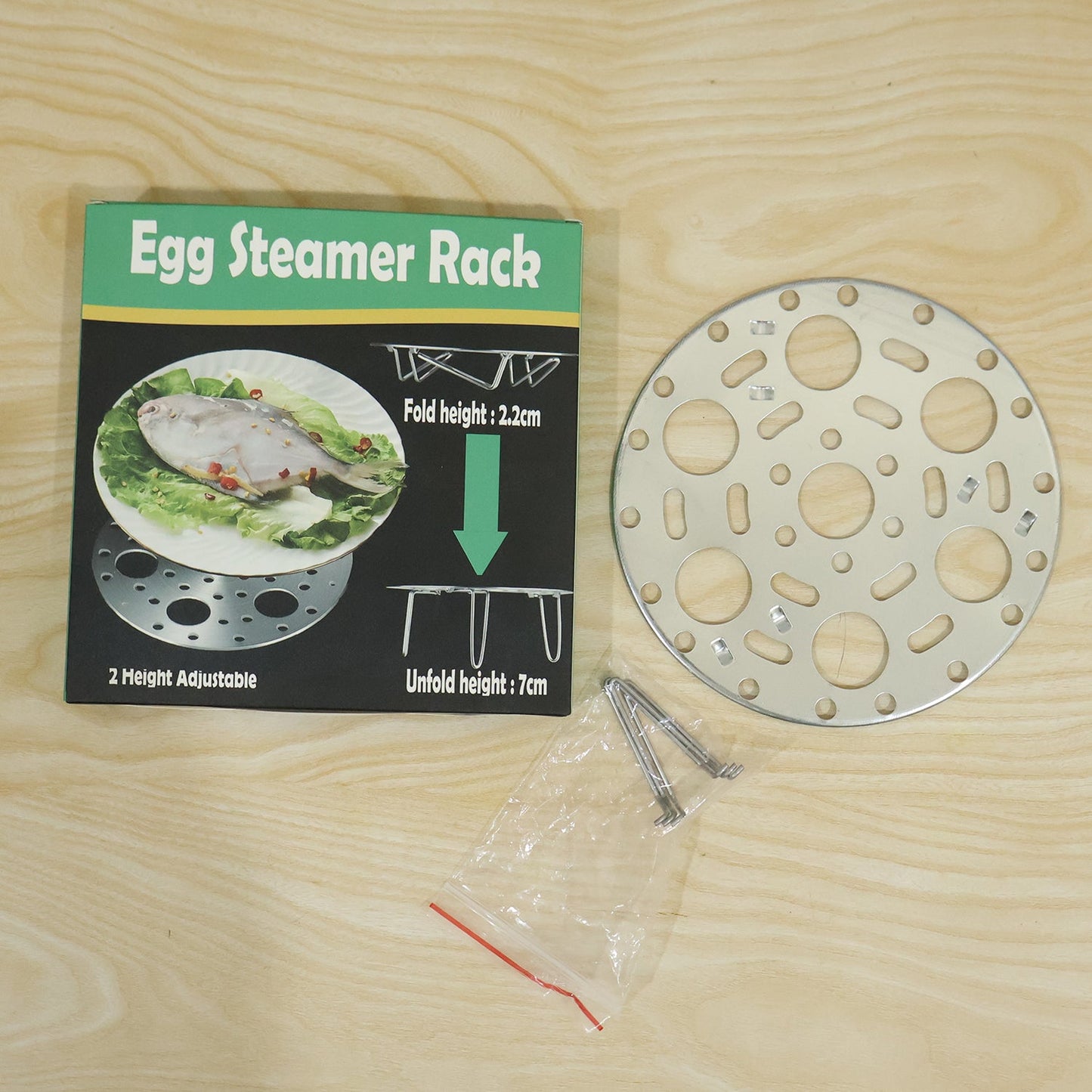 A stainless steel egg steamer rack, its packaging box, and a small plastic bag of extra legs are displayed on a light wooden