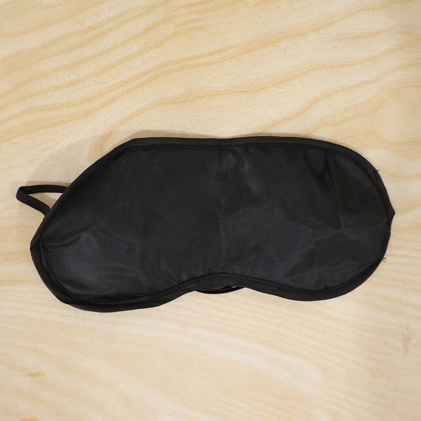 A black silk satin sleep mask with elastic straps lying flat on a light wooden surface.