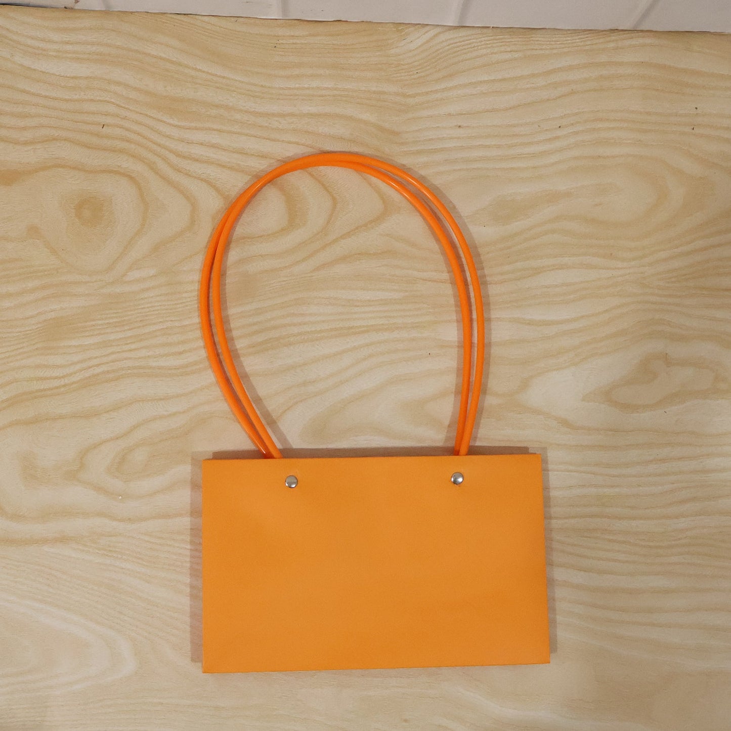 A glossy orange rectangular gift bag with thin, round orange handles is centered on a light wooden background.