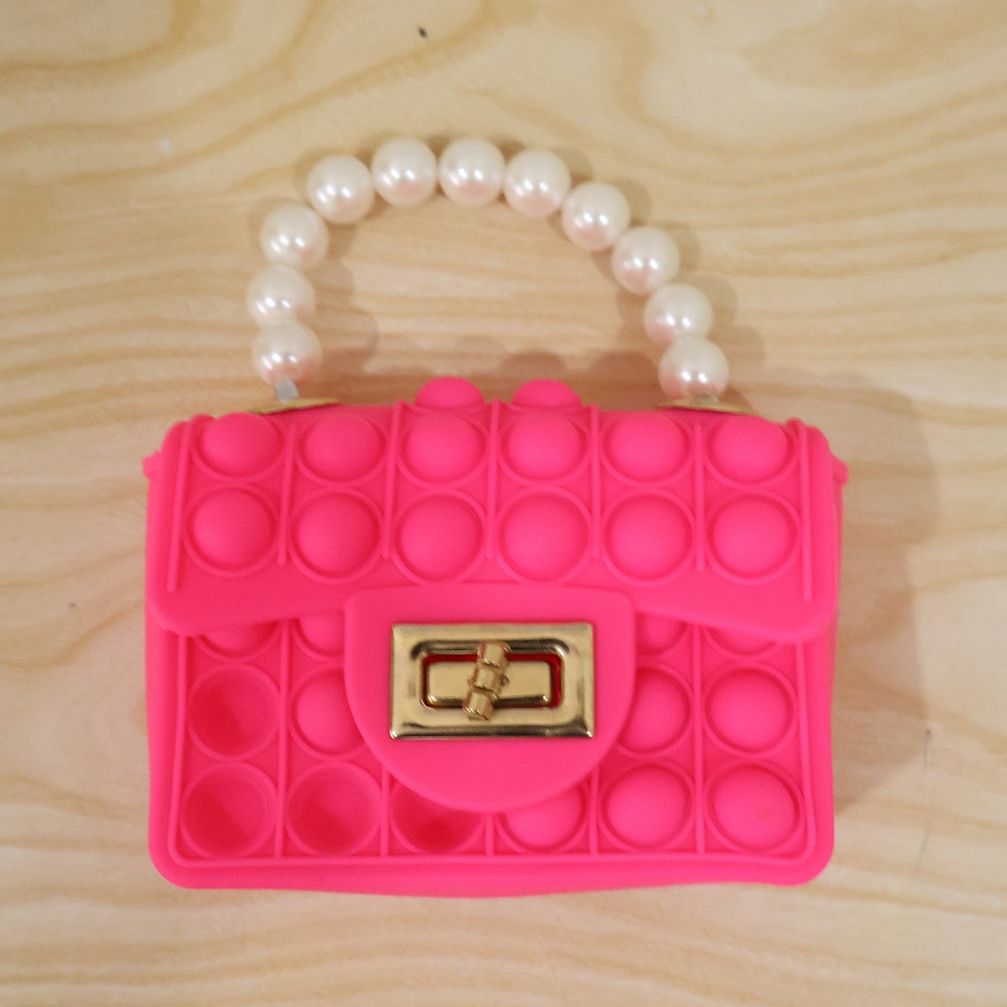 A bright pink pop-it purse with a pearl handle and gold clasp is displayed on a light wooden surface.