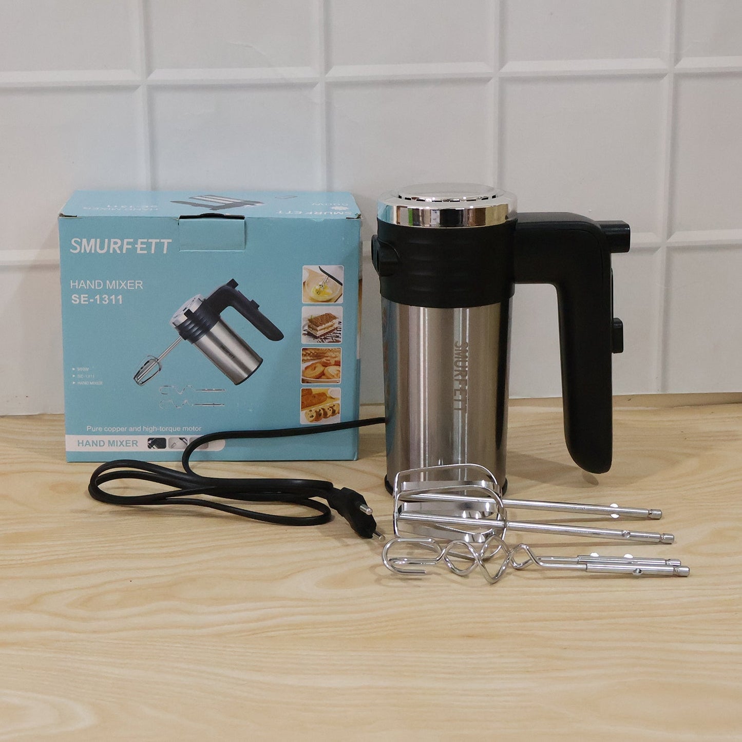 A light blue product box and a black and silver electric hand mixer with beaters and dough hooks are on a light wooden