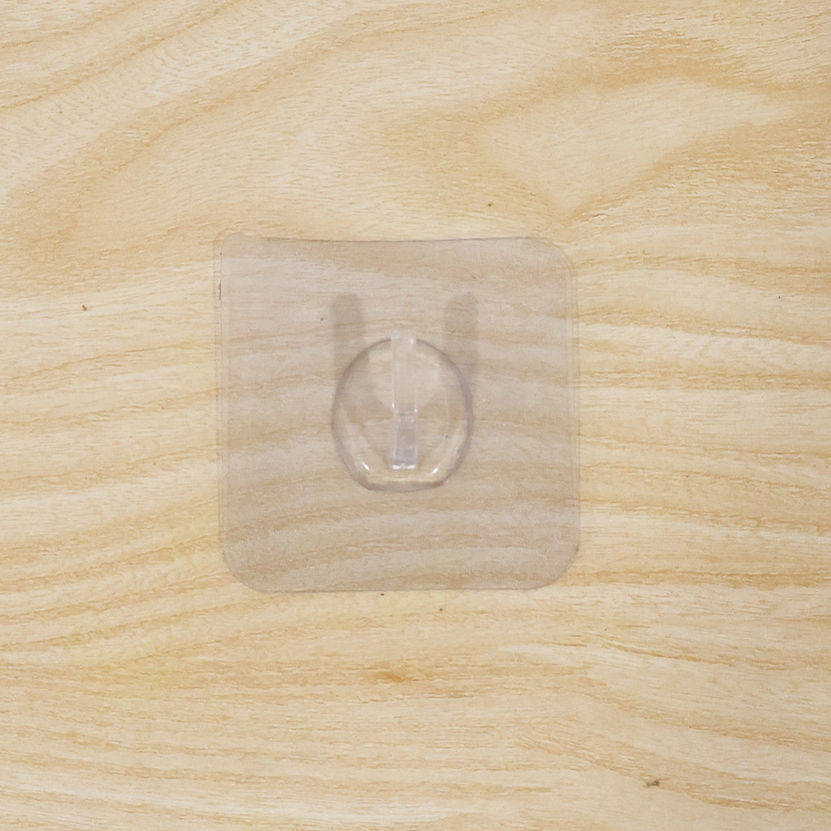 A clear self-adhesive wall hook centered on a light, unfinished wooden surface with a visible grain.
