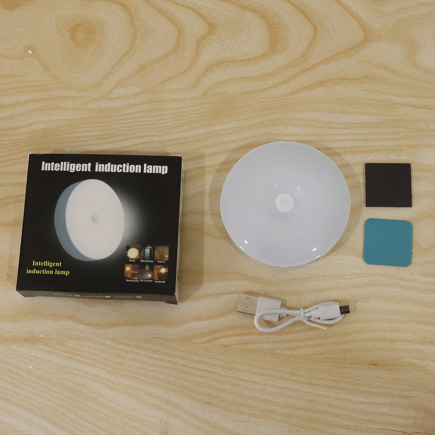 A black box labeled "Intelligent induction lamp," a white round LED light, a USB cable, and two adhesive pads on a wooden