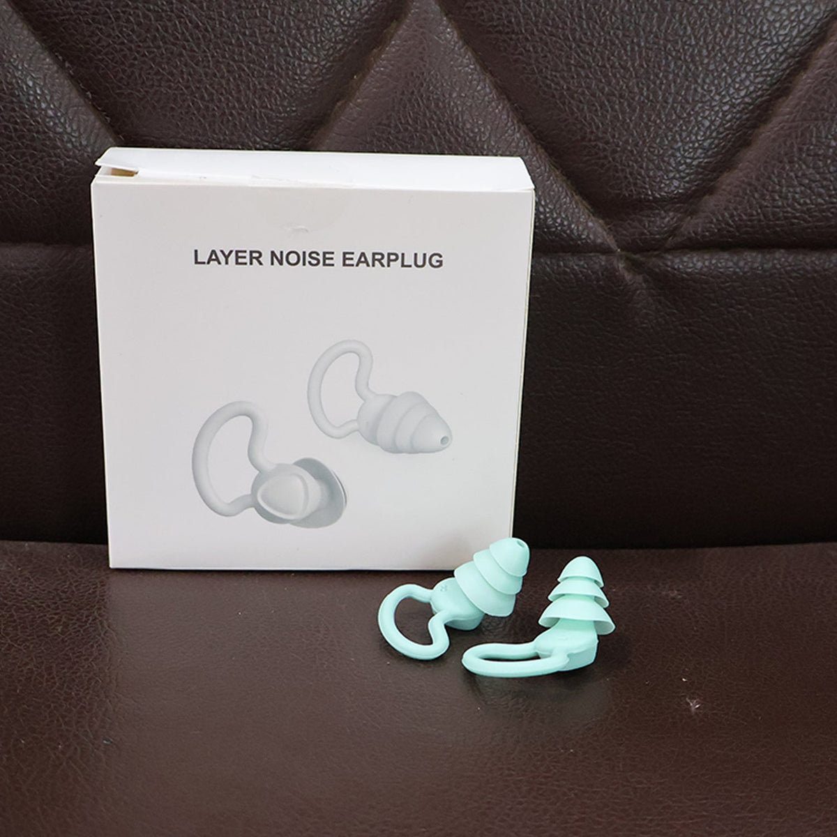 A white box labeled 'LAYER NOISE EARPLUG' sits next to a pair of mint green silicone earplugs on a brown surface.