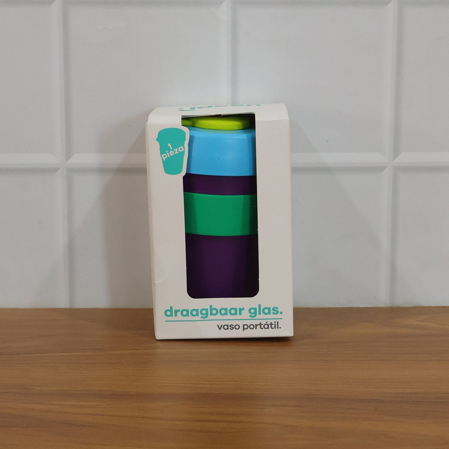 A white box containing a purple, green, and blue reusable cup, labeled "draagbaar glas. vaso portátil" on a wooden surface.