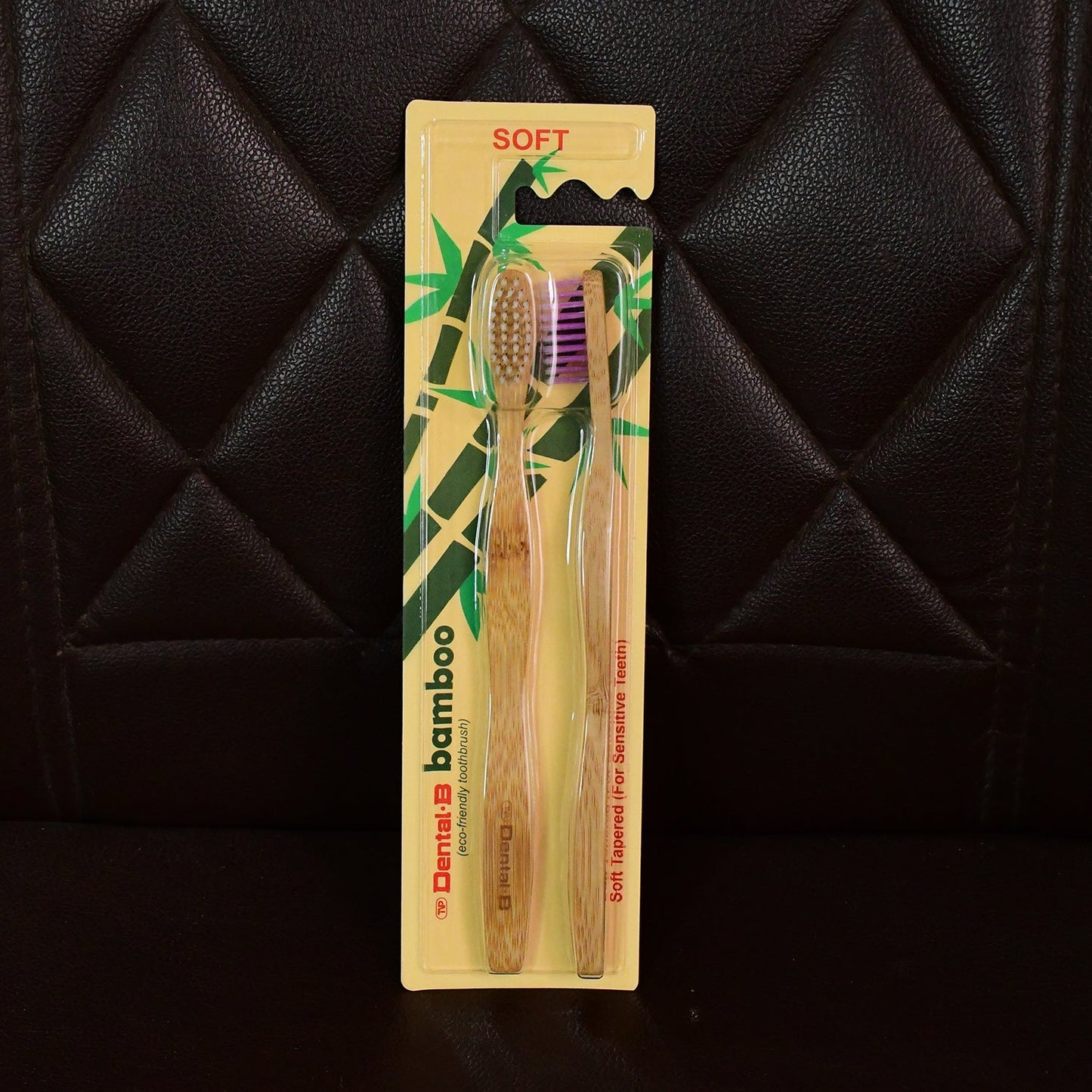 A two-pack of Dental B bamboo toothbrushes, one with brown and one with purple bristles, in yellow packaging against a dark