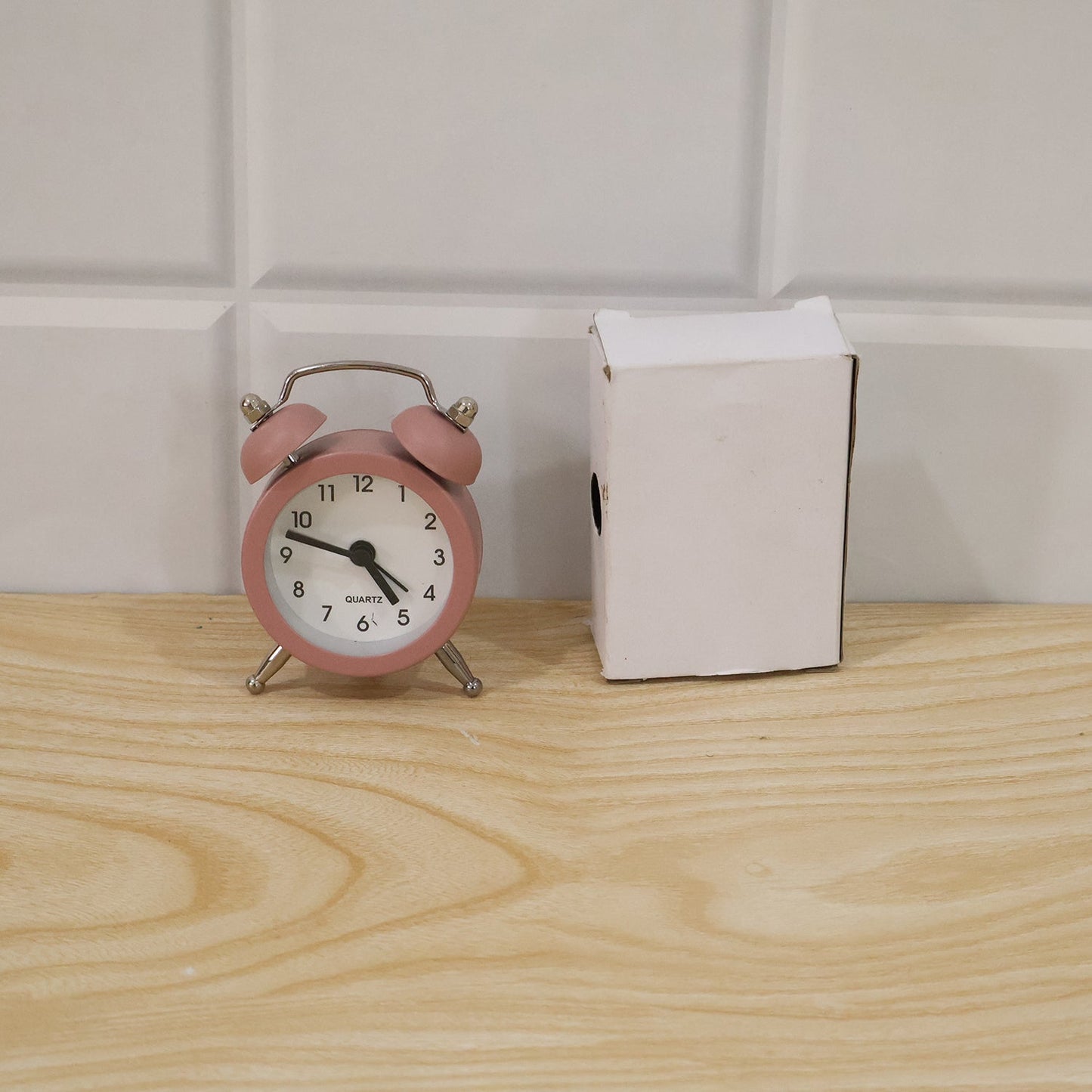 Pink twin bell alarm clock next to a small white open box on a light wooden surface with white tiled wall.