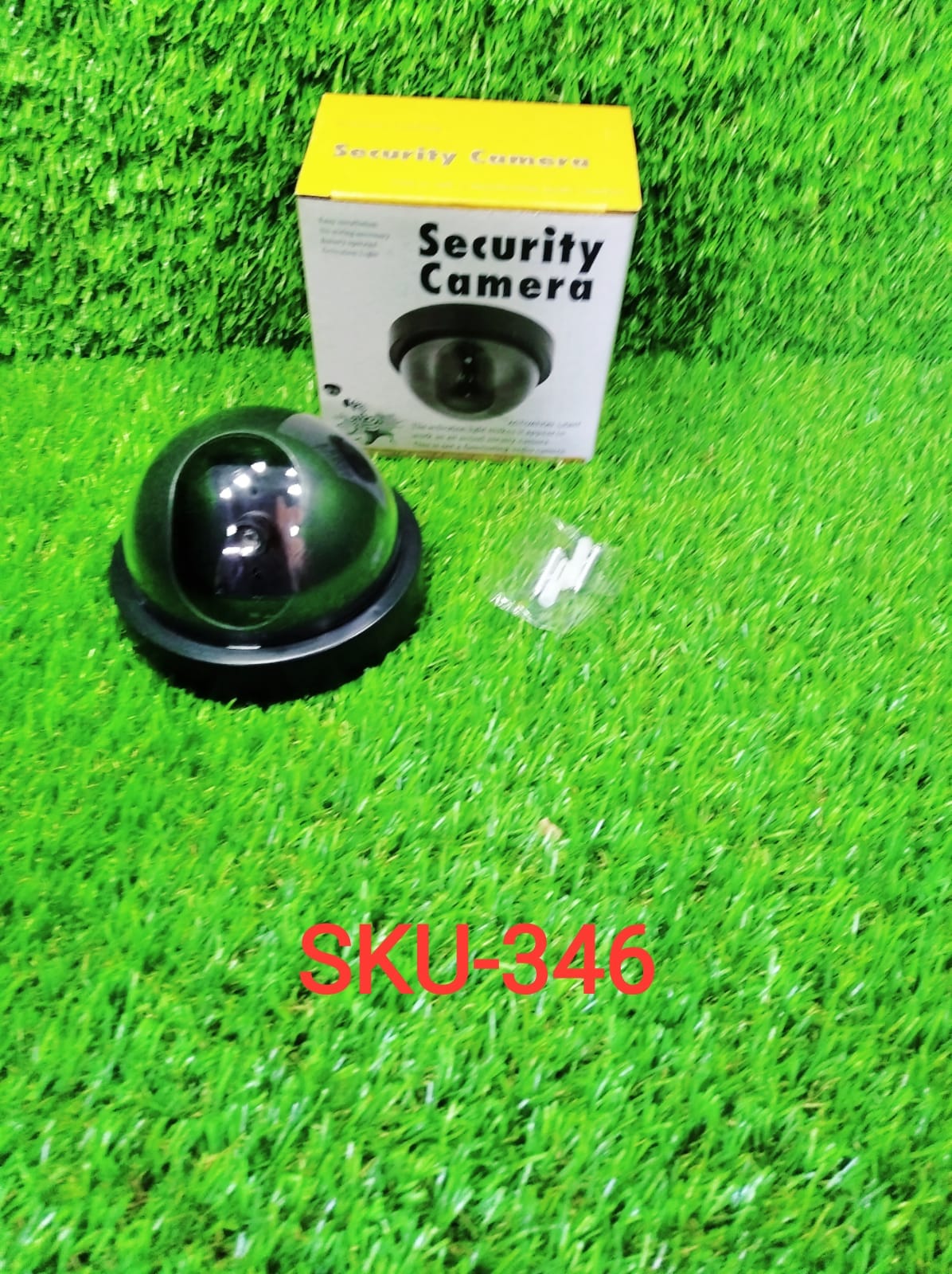 Black dome dummy security camera next to its yellow and white packaging box and small white screws, all on artificial green