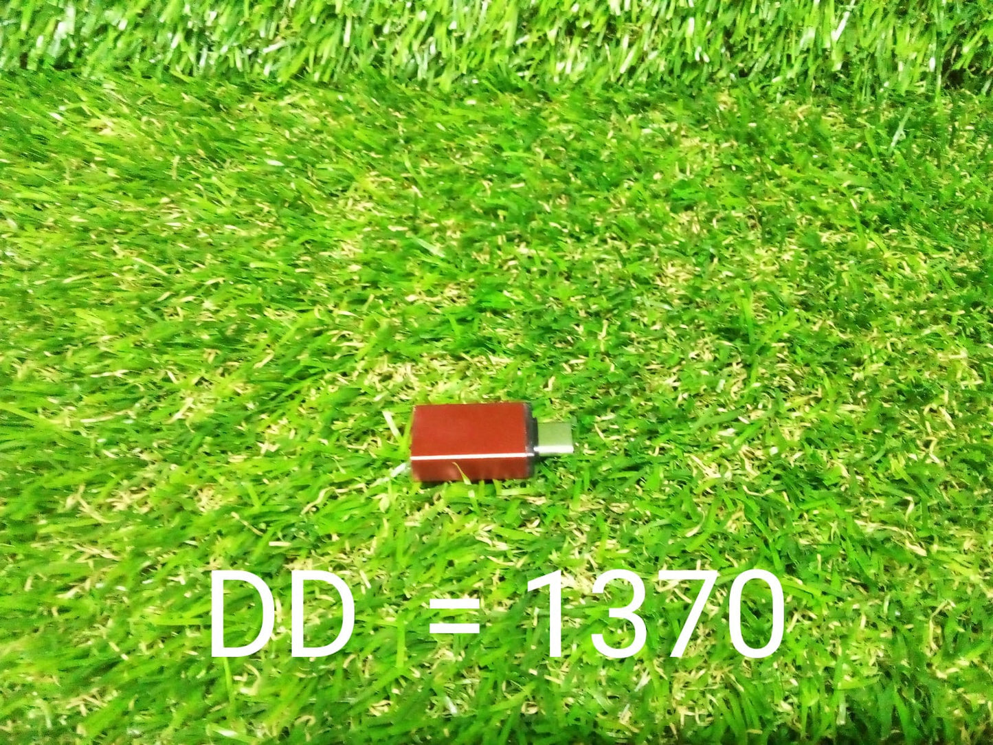 A small red USB-C to USB 3.0 OTG adapter is centered on a vibrant green artificial grass background.