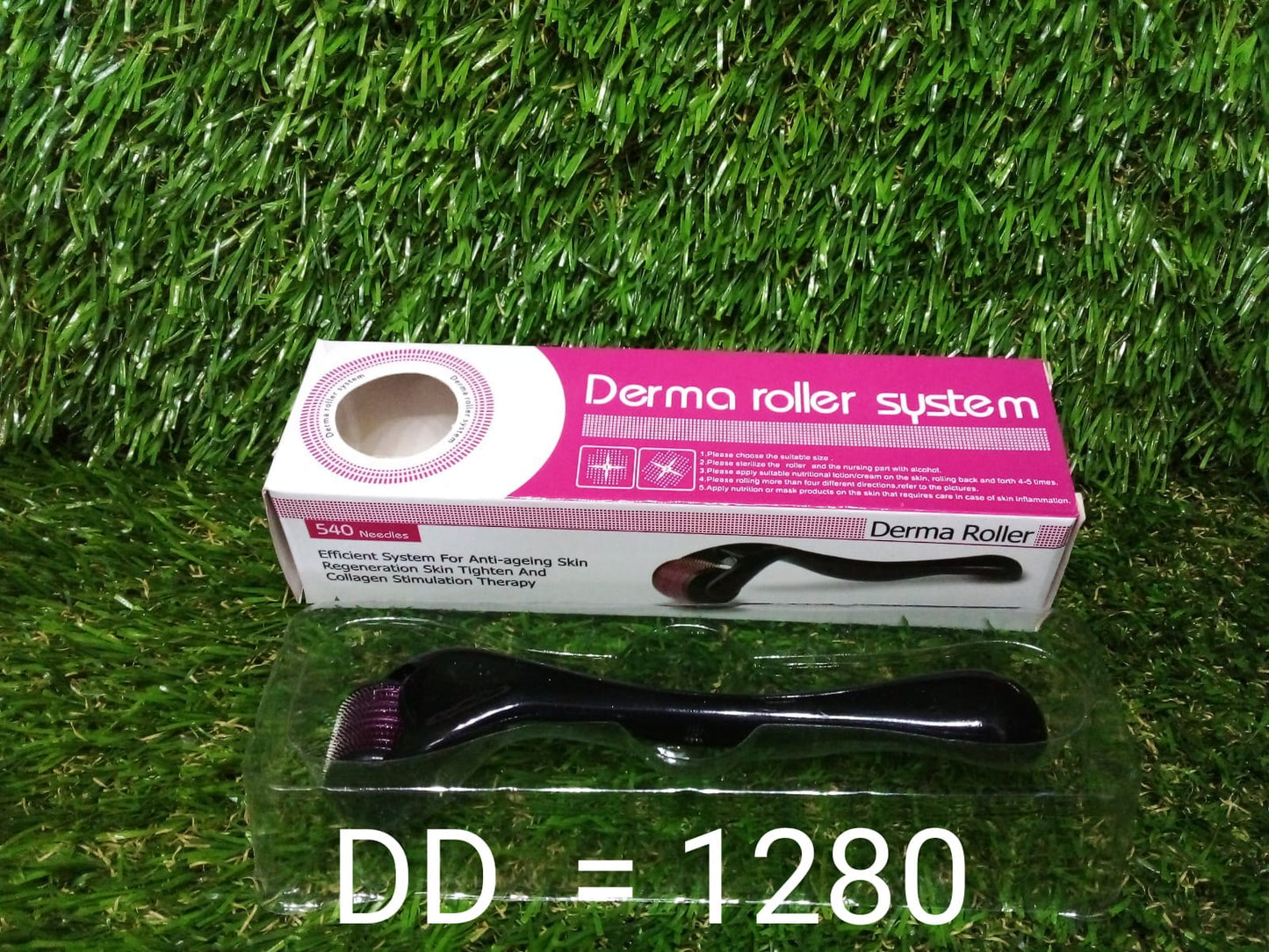 A derma roller in a clear plastic tray and its pink and white box are displayed on artificial grass.