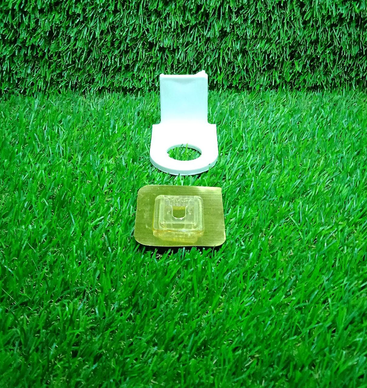 A white plastic bottle holder and a metallic adhesive pad are displayed separately on a vibrant green artificial grass