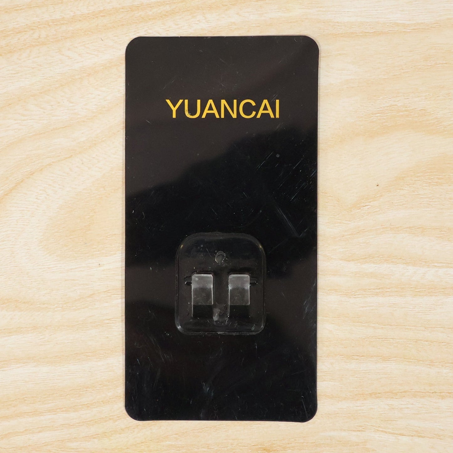A black rectangular adhesive wall hook with gold text and a clear plastic hook on a light wooden surface.