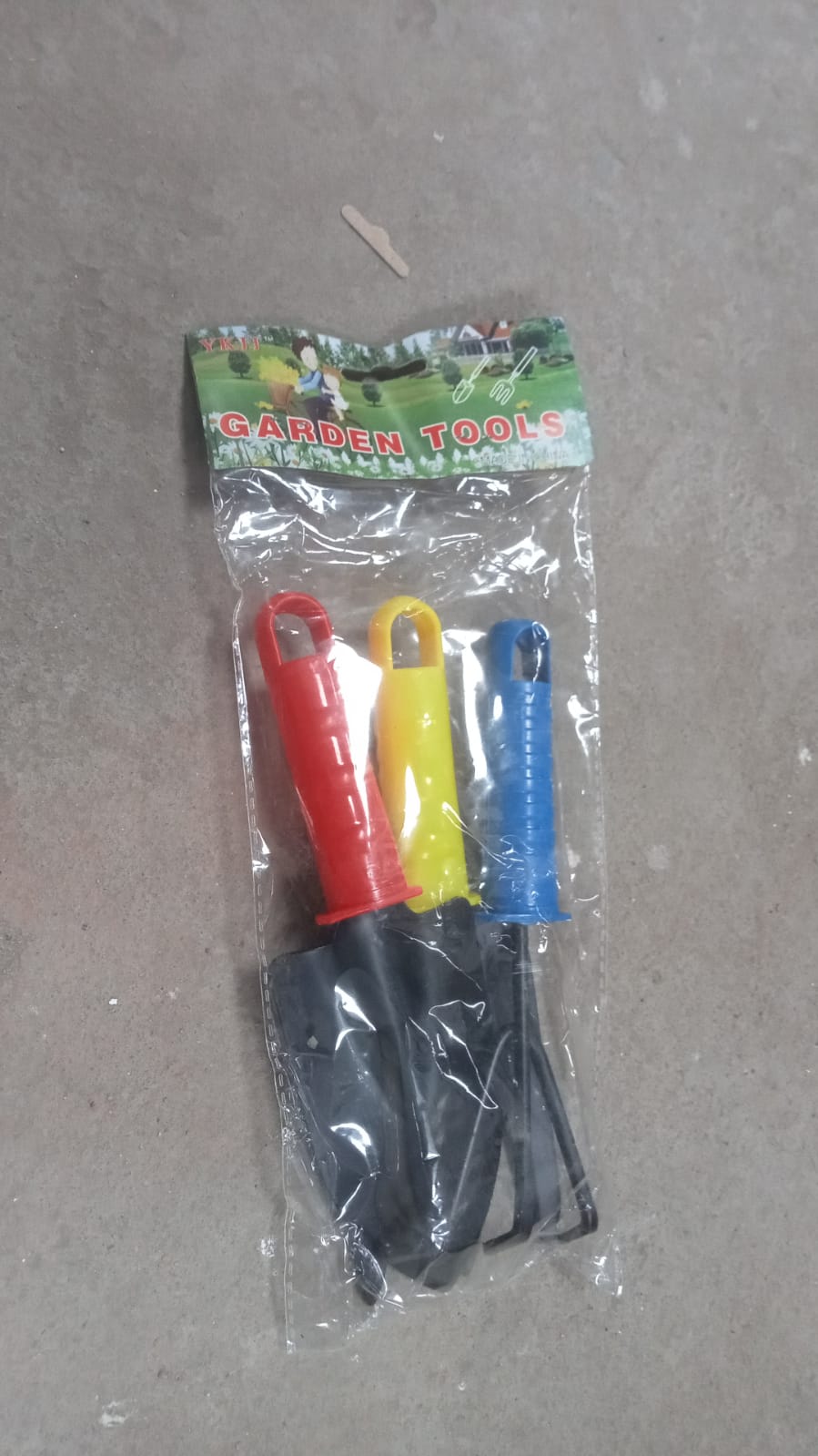 A clear plastic package contains a red, yellow, and blue handled garden tool set, lying on a concrete surface.