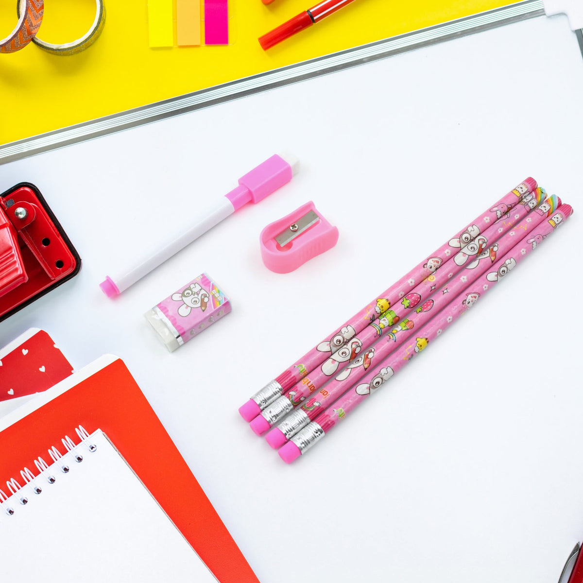 Pink bunny pencils, a marker, an eraser, and a sharpener are arranged on a white surface with red and yellow office supplies.