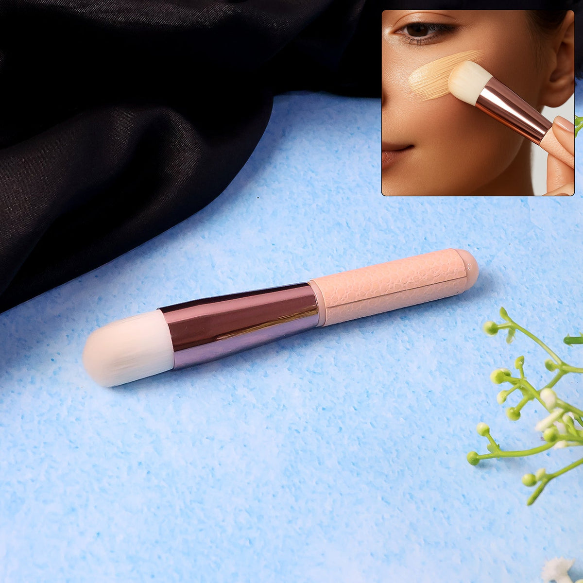 A pink and rose gold makeup brush on a blue textured surface with a black fabric and green leaves.