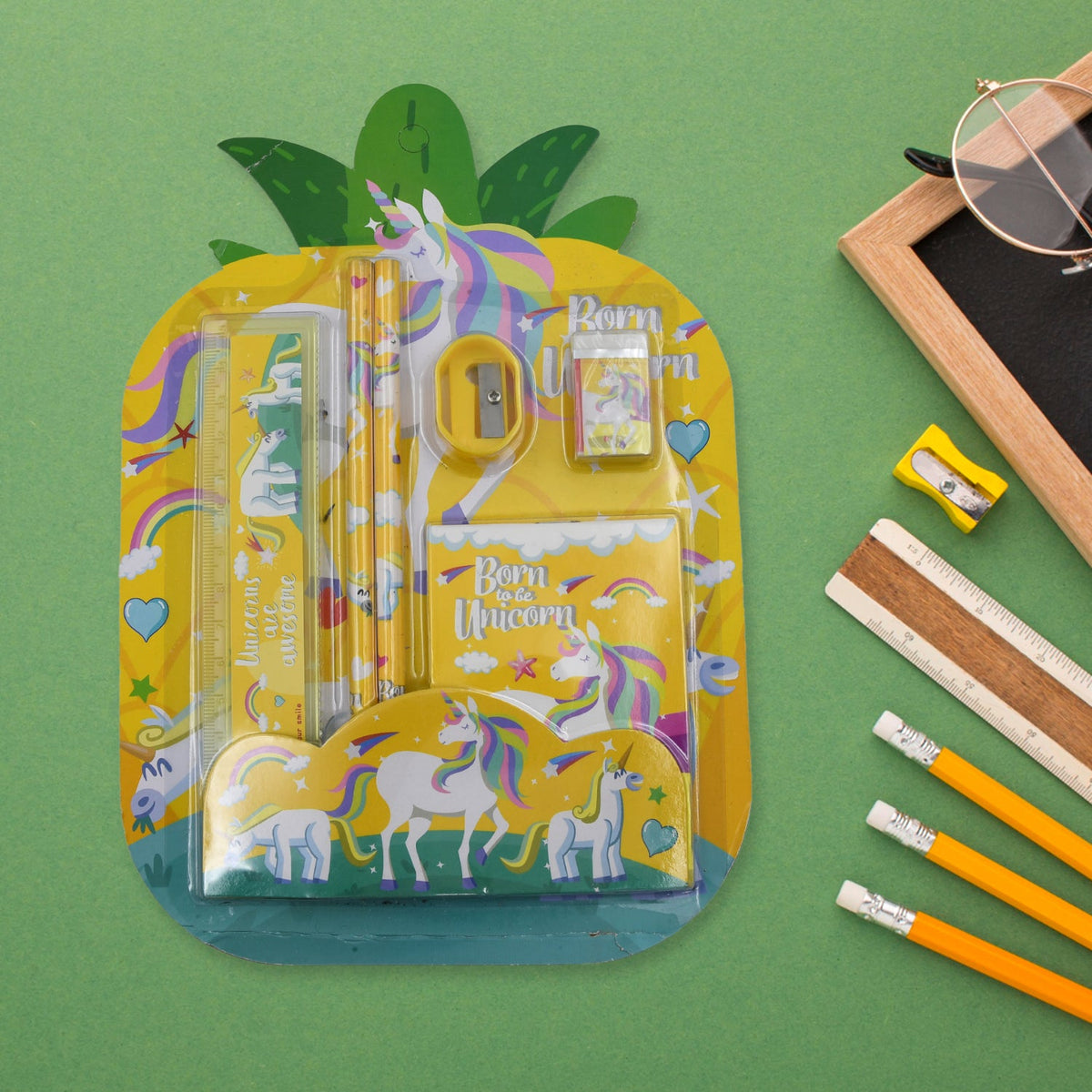 A yellow unicorn stationery set in pineapple packaging on a green background, with a small chalkboard, glasses, and pencils.