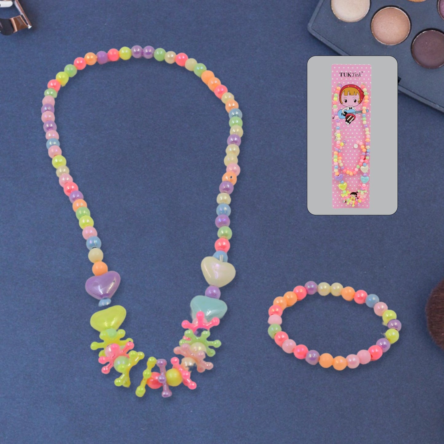 A colorful beaded necklace and bracelet set is displayed on a dark blue surface, with the pink packaging and an eyeshadow
