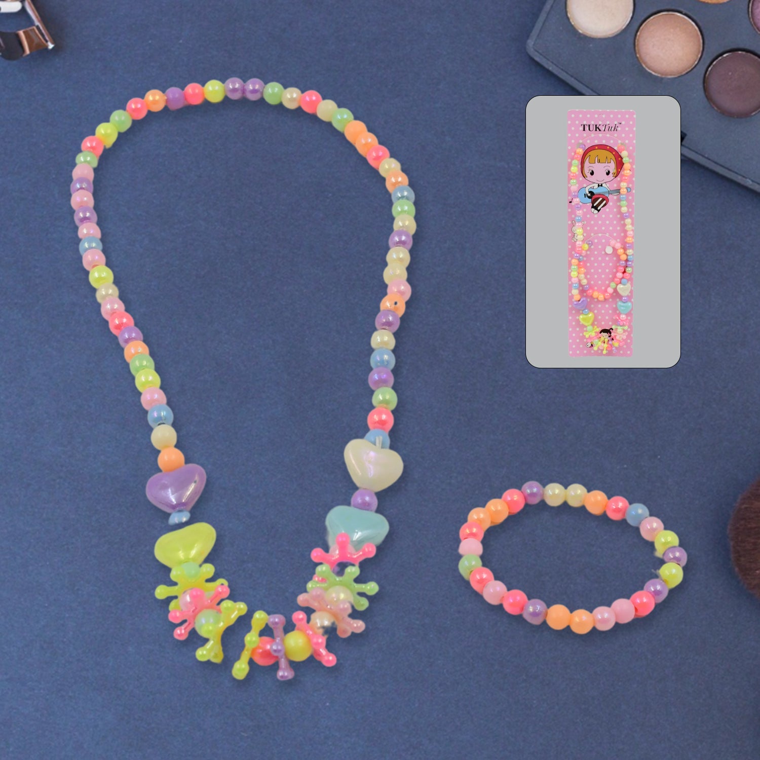 A colorful beaded necklace and bracelet set is displayed on a dark blue surface, with the pink packaging and an eyeshadow