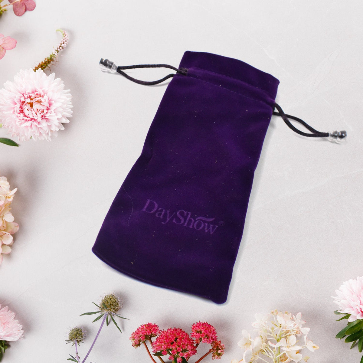 A purple velvet drawstring bag with a DayShow logo is centered on a white surface with various pink and white flowers.