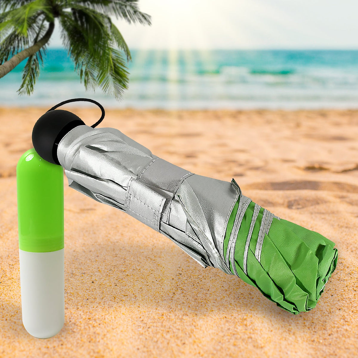 A compact green and silver umbrella, rolled up, rests on a sandy beach next to its green and white capsule case.