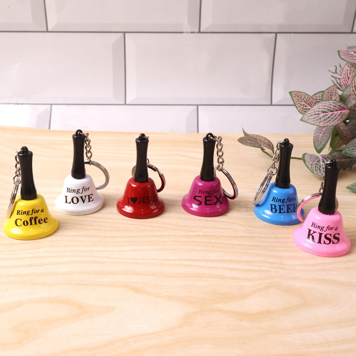 Six mini hand bell keychains in various colors and phrases, displayed on a wooden surface with a white tiled wall in the