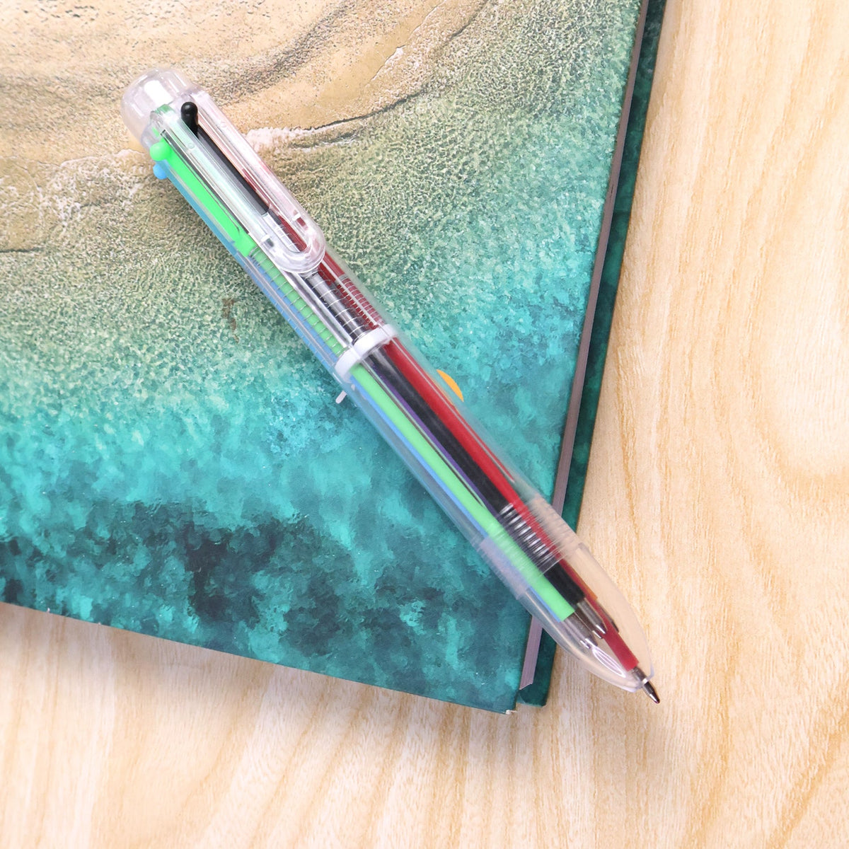 A clear 6-in-1 multicolor ballpoint pen with red, black, green, and blue ink cartridges rests on a teal and brown surface.