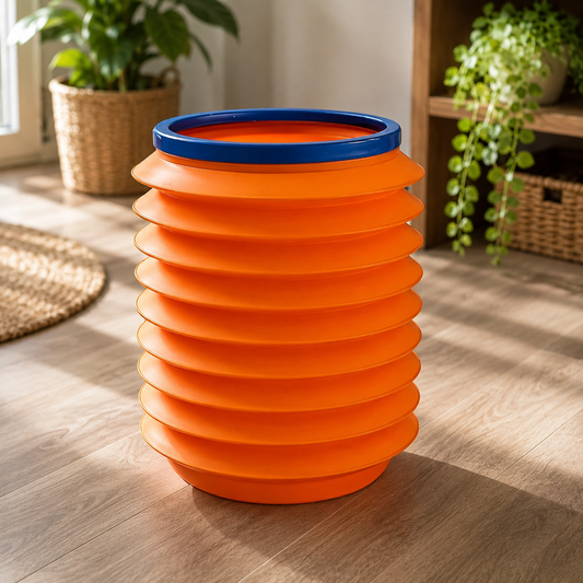An orange accordion-style storage bucket with a blue rim sits on a wooden floor in a room with plants.