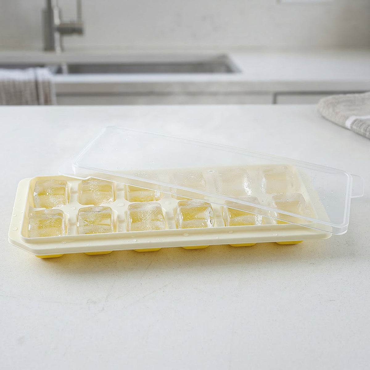 A yellow silicone ice cube tray with a clear lid partially open, filled with ice cubes, sits on a white kitchen counter.