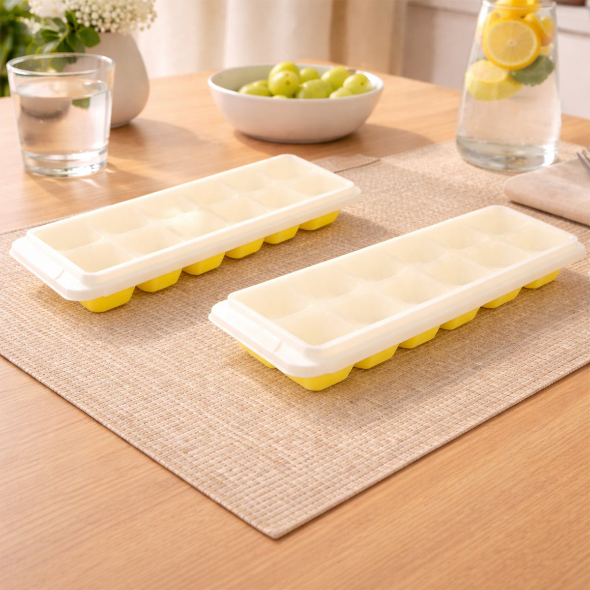 Two yellow silicone ice cube trays with clear lids sit on a woven placemat on a wooden table with drinks and fruit.